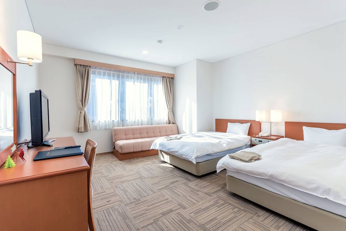 A hotel room with a bed, a desk with a computer, a chair, a sofa, and a window with curtains.