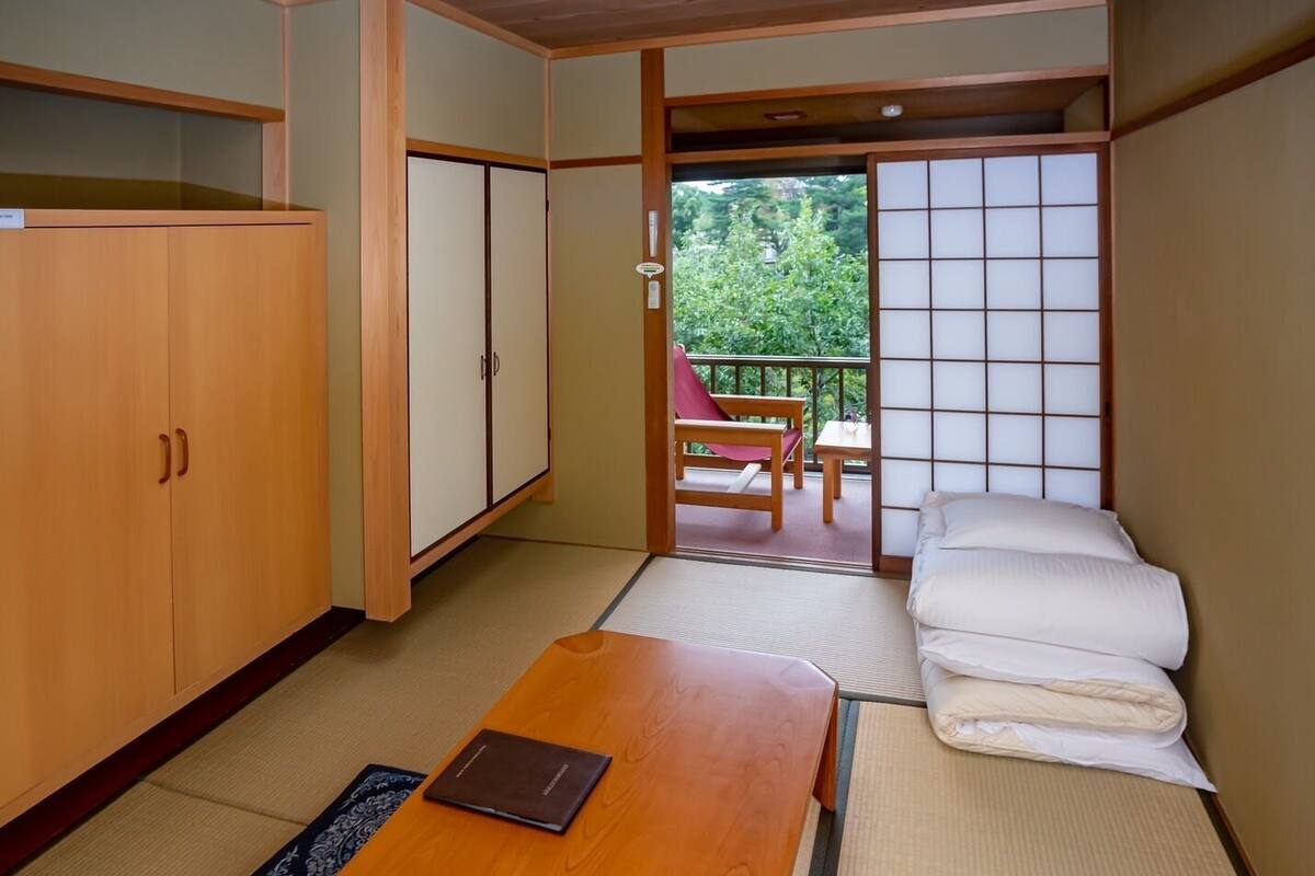 A traditional Japanese room with tatami flooring, a low wooden table, a futon bed, and sliding doors leading to an outdoor balcony.