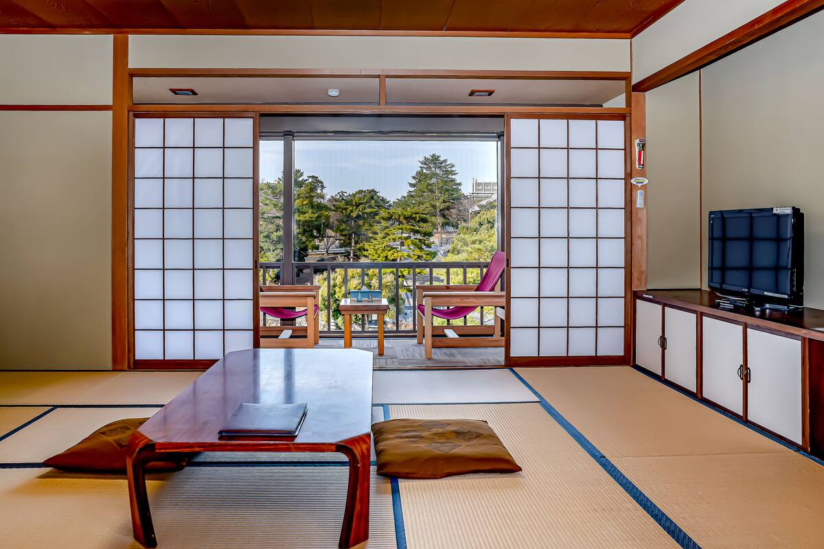 A traditional Japanese room with tatami flooring, a low table, cushions, a TV, and a view of trees and buildings outside.