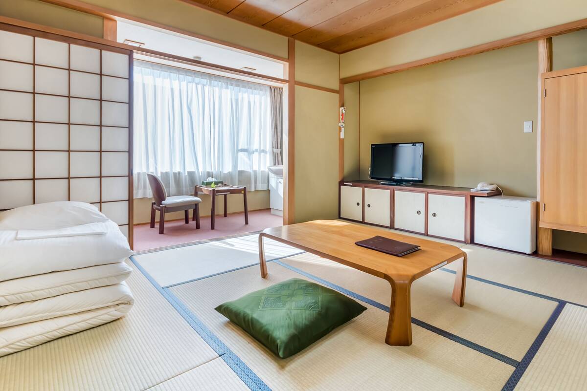 A traditional Japanese-style room with tatami flooring, a low wooden table, a television on a cabinet, and a futon bed.