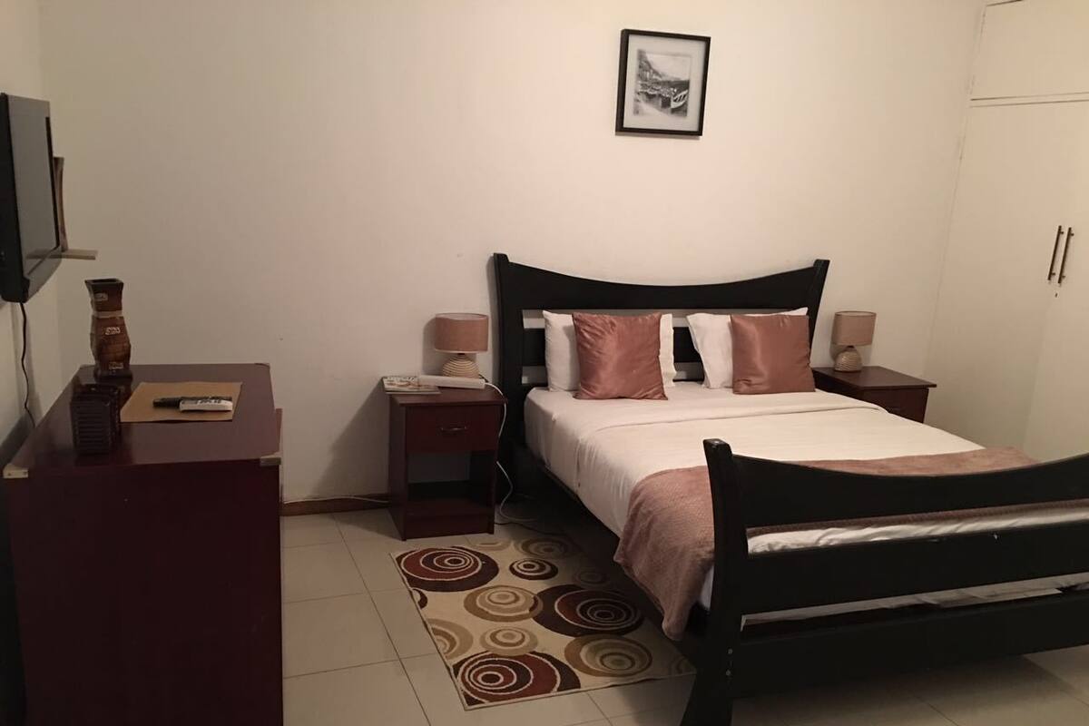 Un dormitorio con cama, escritorio, mesita de noche, lámpara y un cuadro en la pared.