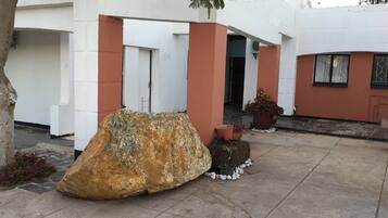 Un patio con una roca grande, plantas en macetas y un edificio blanco con detalles rojos.