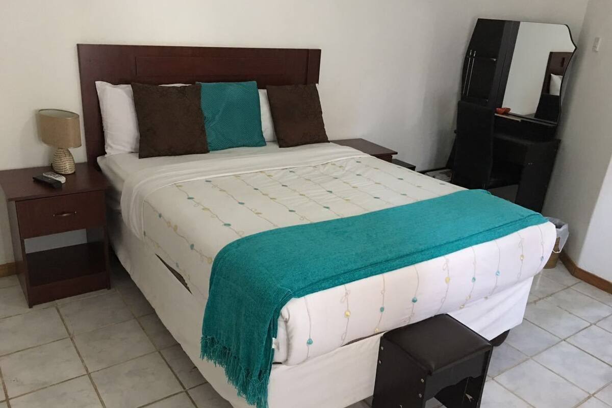 Una cama bien hecha con una manta color turquesa, un cabecero de madera y una mesita de noche con una lámpara y un control remoto.