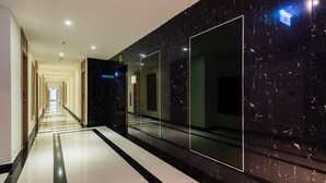 A modern hotel corridor with a reflective black marble wall, a glass elevator, and a clean, tiled floor.