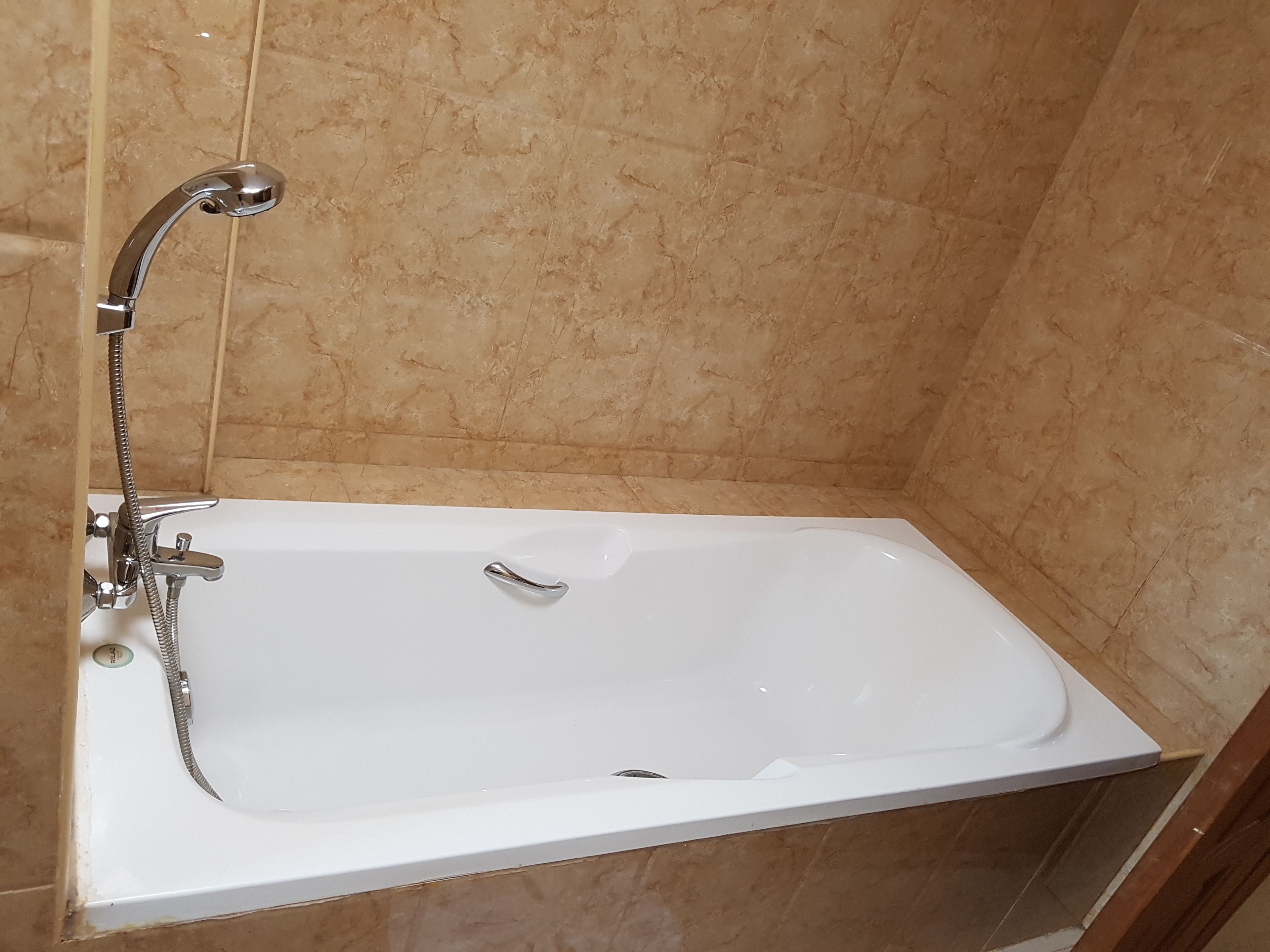 Une baignoire blanche, dotée d’un pommeau de douche à main et d’un robinet séparé, le tout installé sur un mur recouvert de carreaux de marbre beige.
