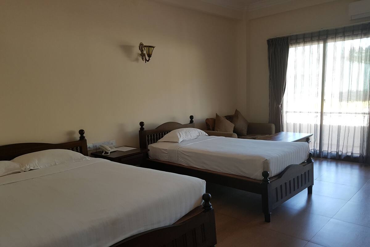 Quarto de hotel com duas camas de solteiro, mesa de cabeceira com telefone, luminária na parede e uma janela grande com cortinas.