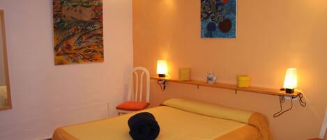 A hotel room with a bed, a chair, a shelf with decorative items, and wall art.