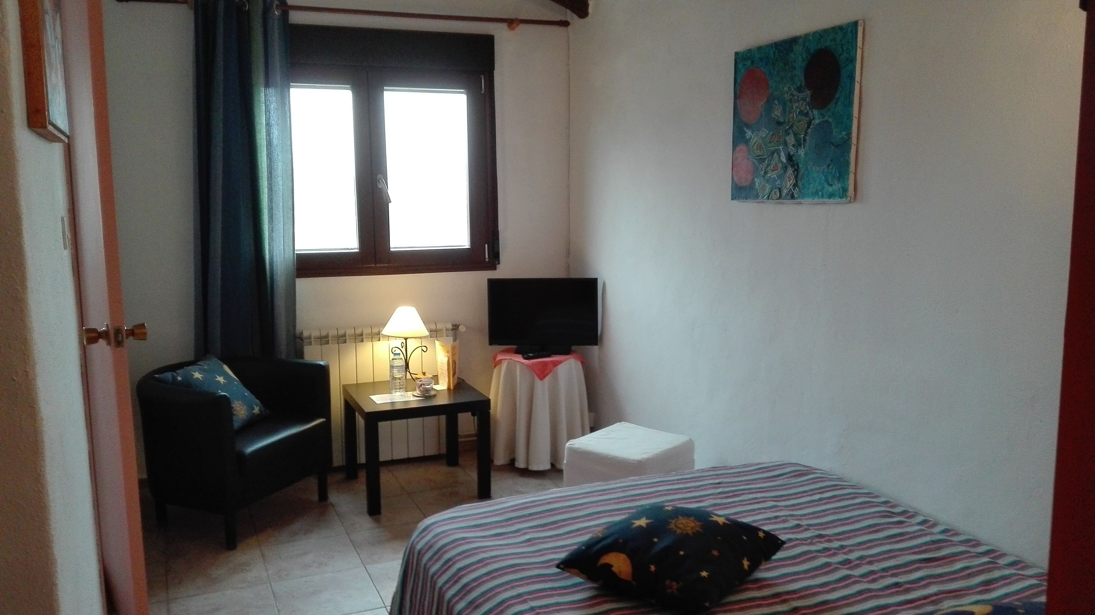 Een compacte, verzorgde kamer met een bed, een stoel, een tafel met een lamp, een televisie op een meubelstuk en een raam met gordijnen.