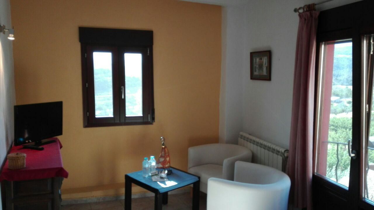 A room with a television on a red table, a small table with a bottle and glasses, two white chairs, a window with a view, and a radiator.
