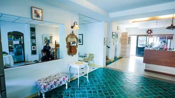 Un vestíbulo de hotel con una mesa de recepción, una zona de estar con un banco, un comedor con una mesa y sillas, y un suelo de baldosas.