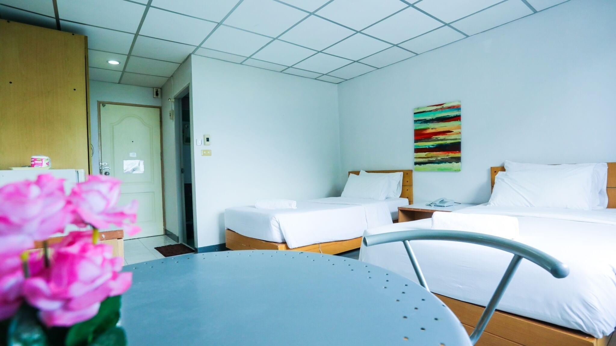 A hotel room with two single beds, a small table, a chair, and a painting on the wall.