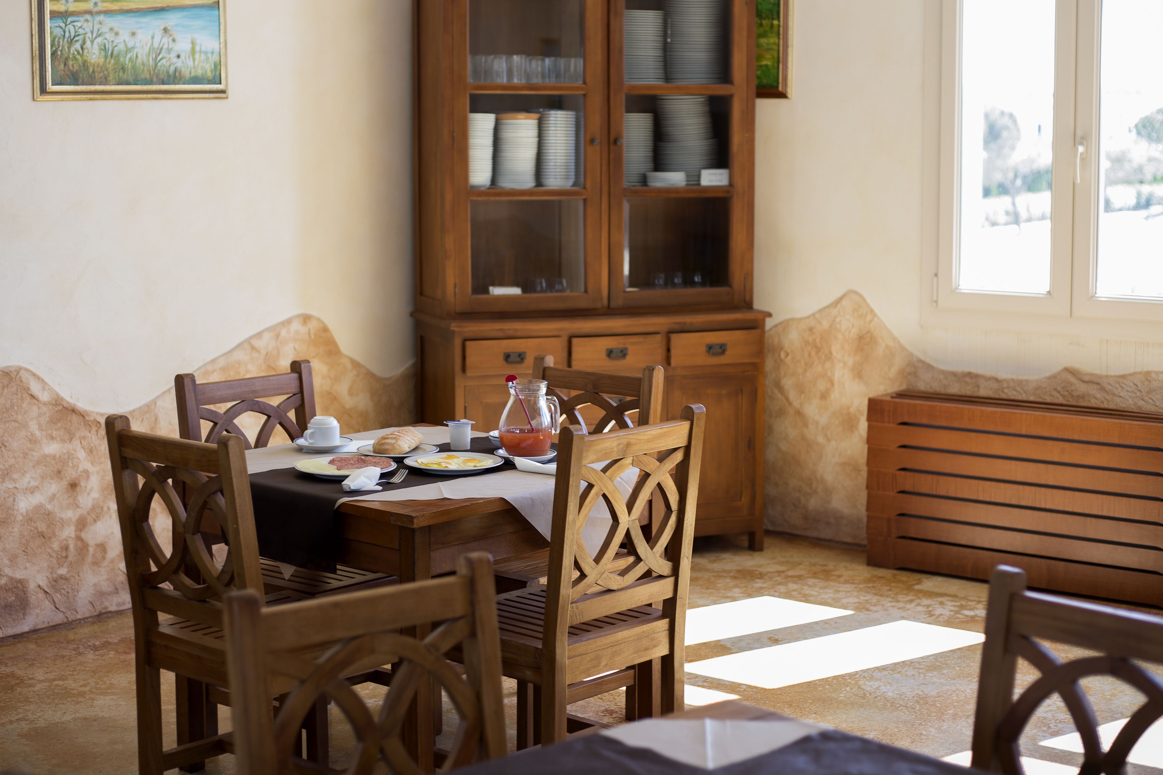 Un coin repas avec des chaises en bois et une table dressée pour quatre personnes, un meuble avec des portes en verre et une vue sur l’extérieur par une fenêtre.