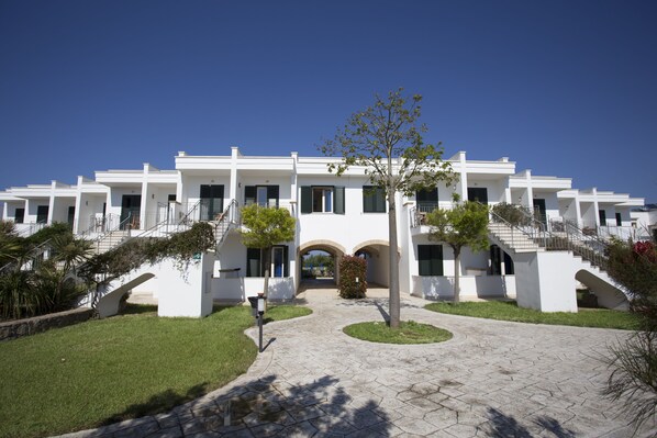 A white building with multiple balconies, a central archway, and a paved courtyard with a tree. - Hotel Residence Portoselvaggio (Nardò)