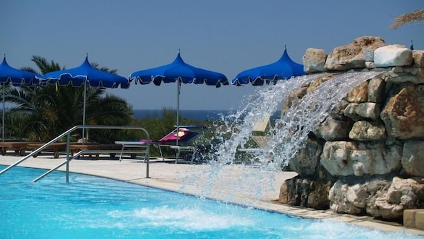 A pool with a waterfall feature, surrounded by blue umbrellas and lounge chairs. - Hotel Residence Portoselvaggio (Nardò)