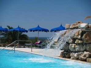 A pool with a waterfall feature, surrounded by blue umbrellas and lounge chairs. - Hotel Residence Portoselvaggio (Nardò)