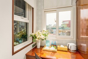 A hotel room with a wooden desk, a vase of flowers, a mini-bar with beverages, and a view of the city through the windows. - Hanoi Maro Hotel (Hanoi)