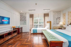 A hotel room with a television, wooden furniture, a bed with a blue and white bedspread, a window with a view of greenery, and decorative wall panels. - Hanoi Maro Hotel (Hanoi)