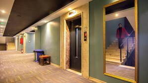 Un couloir d’hôtel avec un ascenseur en marbre, un banc en bois et un grand miroir qui reflète un escalier.