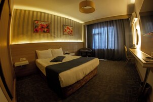 Habitación de hotel con una cama grande, un escritorio, una silla, una lámpara y dos cuadros en la pared.