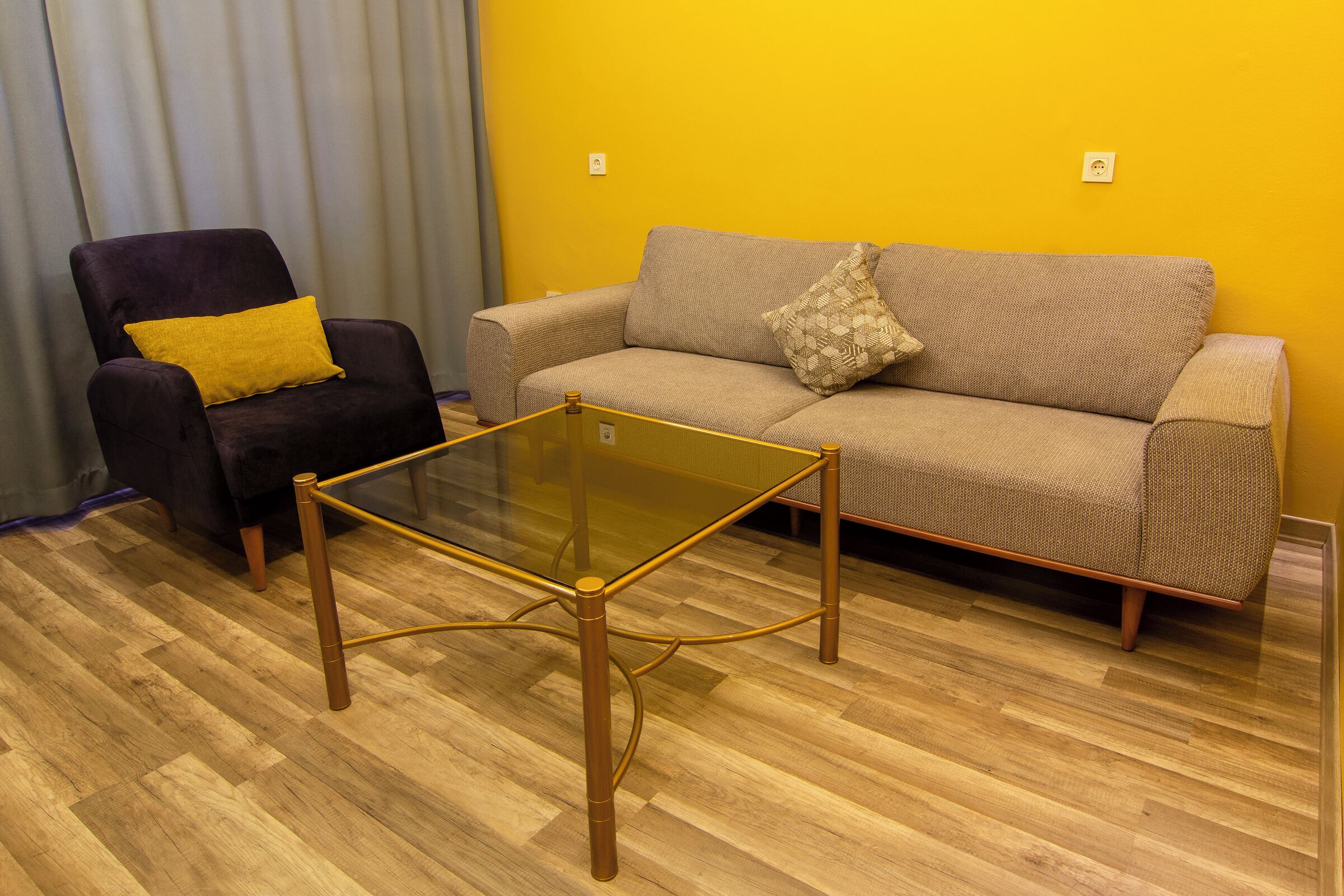 Una sala de estar con una mesa de centro de vidrio, un sillón oscuro con un cojín amarillo y un sofá beige con un cojín estampado.