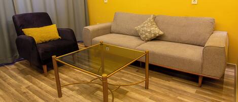 Una sala de estar con una mesa de centro de vidrio, un sillón oscuro con un cojín amarillo y un sofá beige con un cojín estampado.