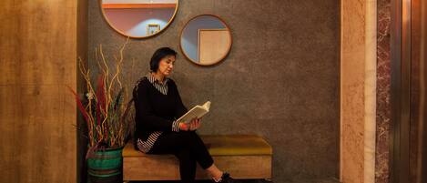 Una persona sentada en un banco leyendo un libro en un vestíbulo con paneles de madera y dos espejos en la pared.
