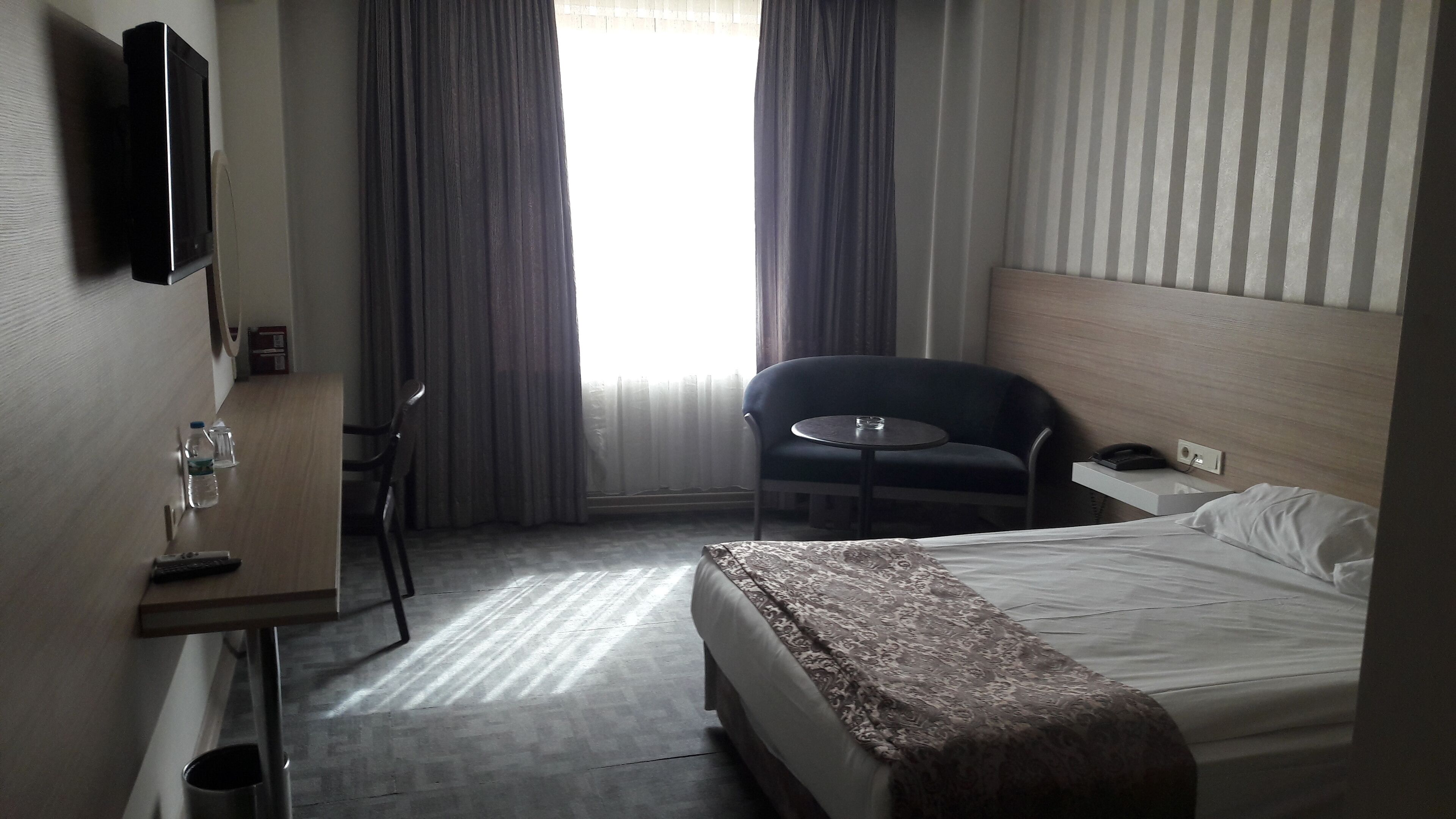 Habitación de hotel con cama, una pequeña mesa con silla, televisor montado en la pared y una ventana con cortinas.