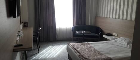Habitación de hotel con cama, una pequeña mesa con silla, televisor montado en la pared y una ventana con cortinas.