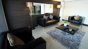 Una sala de estar moderna con paneles de madera oscura, dos sofás negros con cojines, una mesa de centro de vidrio y un espejo grande que refleja la habitación.