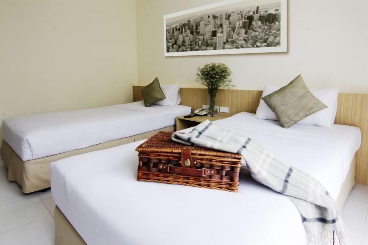 Una habitación de hotel con dos camas individuales, una mesita de noche con un teléfono, un jarrón con flores y una fotografía enmarcada de un paisaje urbano en la pared.