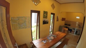 A dining area with a wooden table and benches, a kitchen in the background, and a map on the wall. - Bali Fab Dive Center - Hostel (Karangasem)