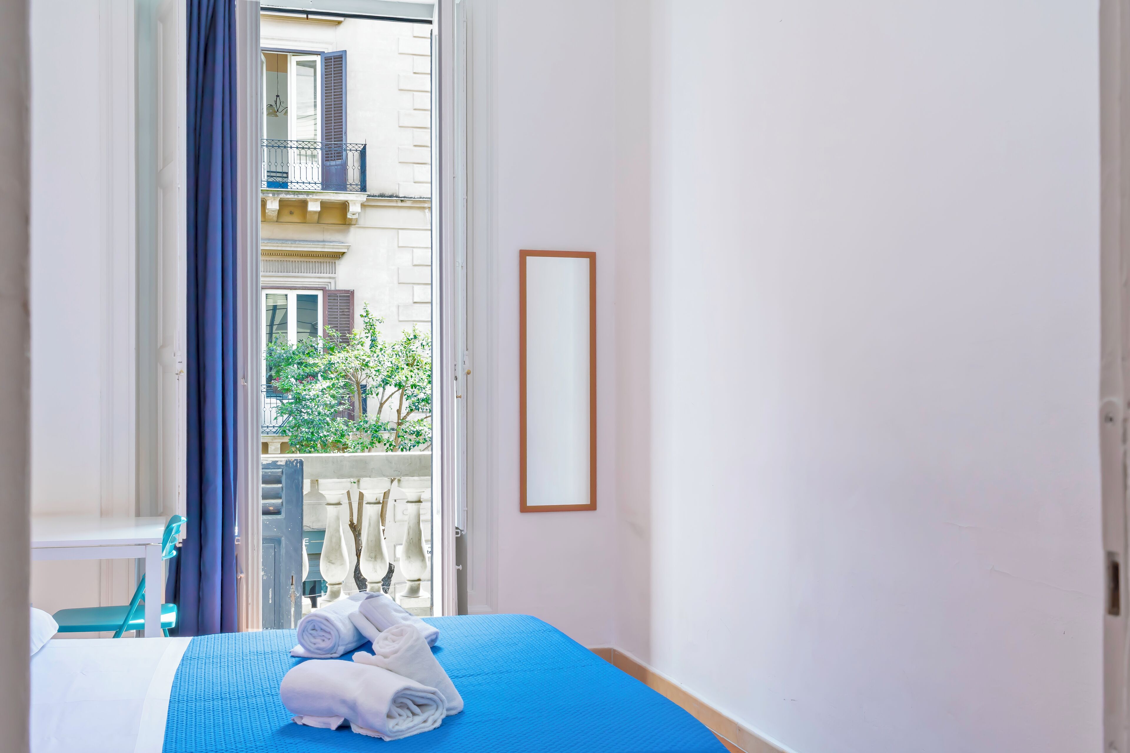 A hotel room with a bed, a window with a view of a balcony and greenery, and a framed picture on the wall.