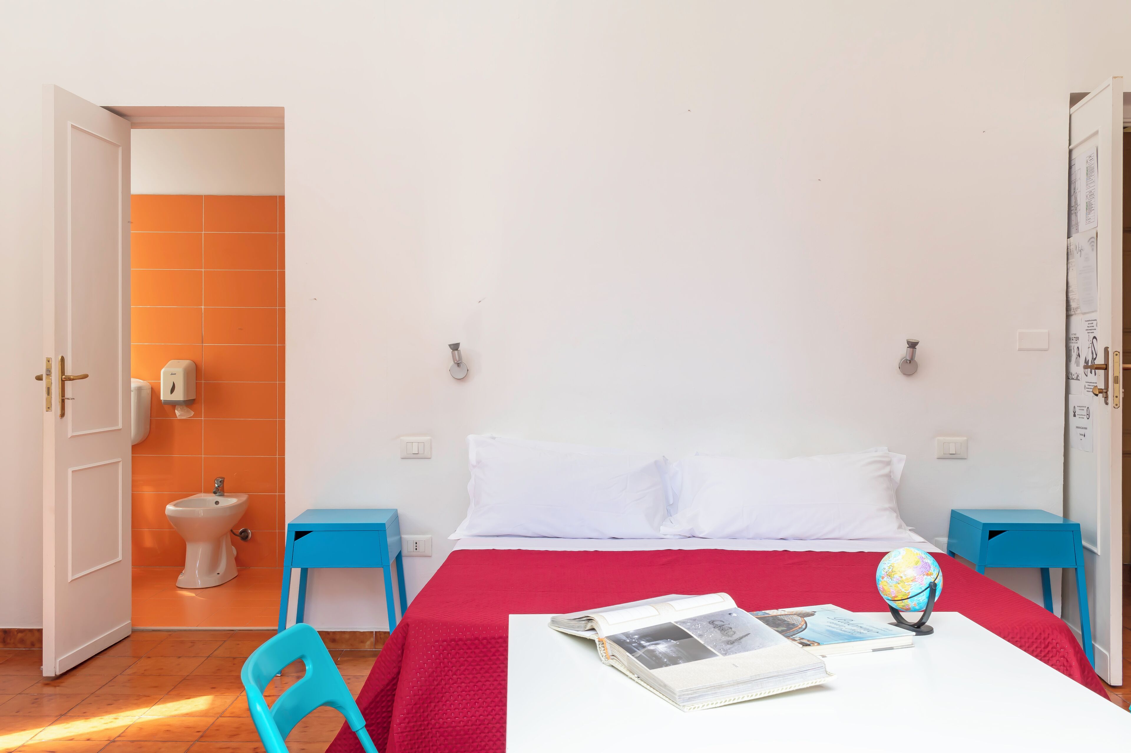 Un dormitorio con una cama, un escritorio con un globo terráqueo y un libro, una silla azul y un baño con pared de azulejos naranjas y un inodoro blanco.