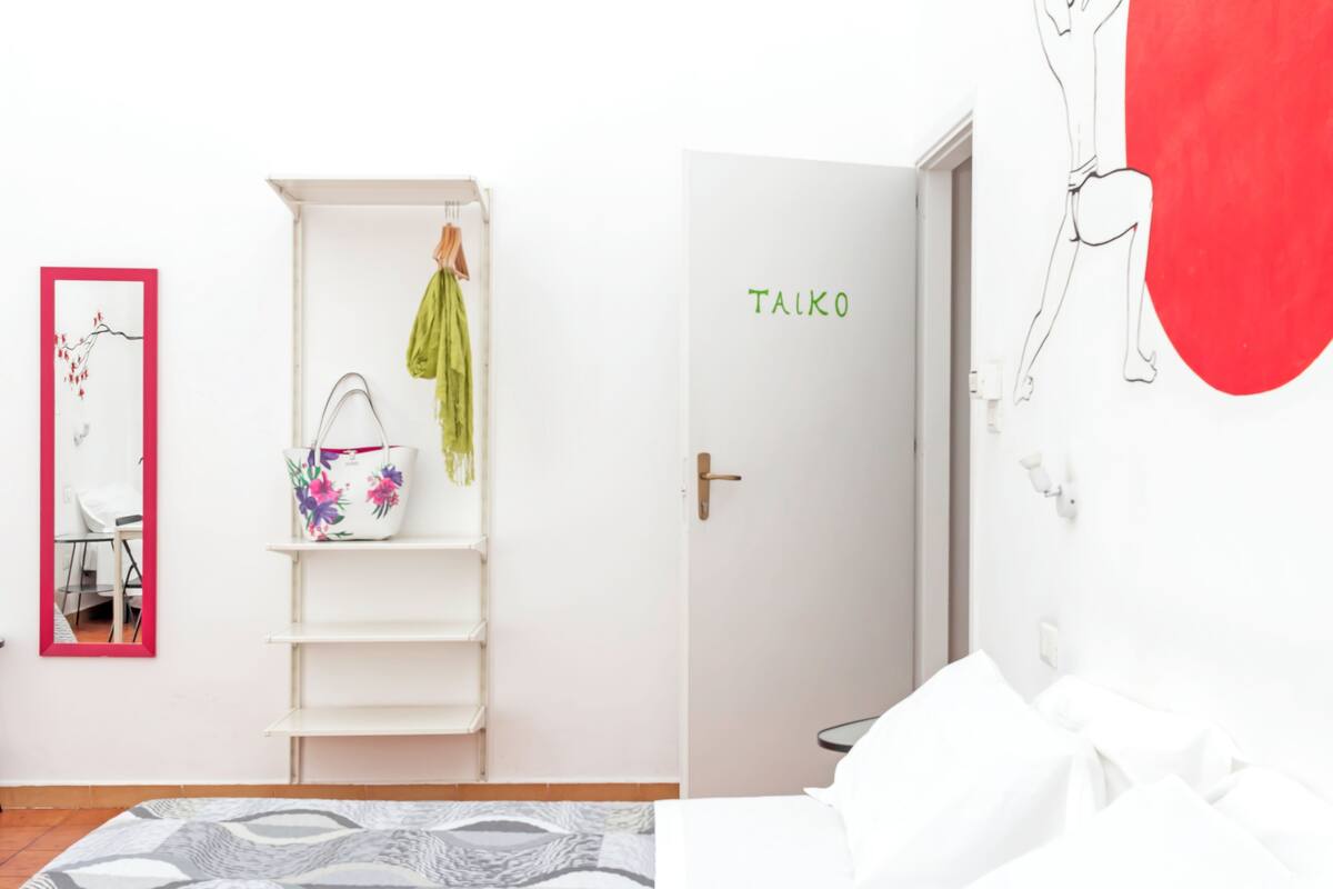 Ein Zimmer mit einer weißen Tür mit der Aufschrift "TALKO", einem weißen Regal mit einem Blumenkorb, einem Spiegel mit rotem Rahmen und einem Bett mit weißer Bettwäsche.