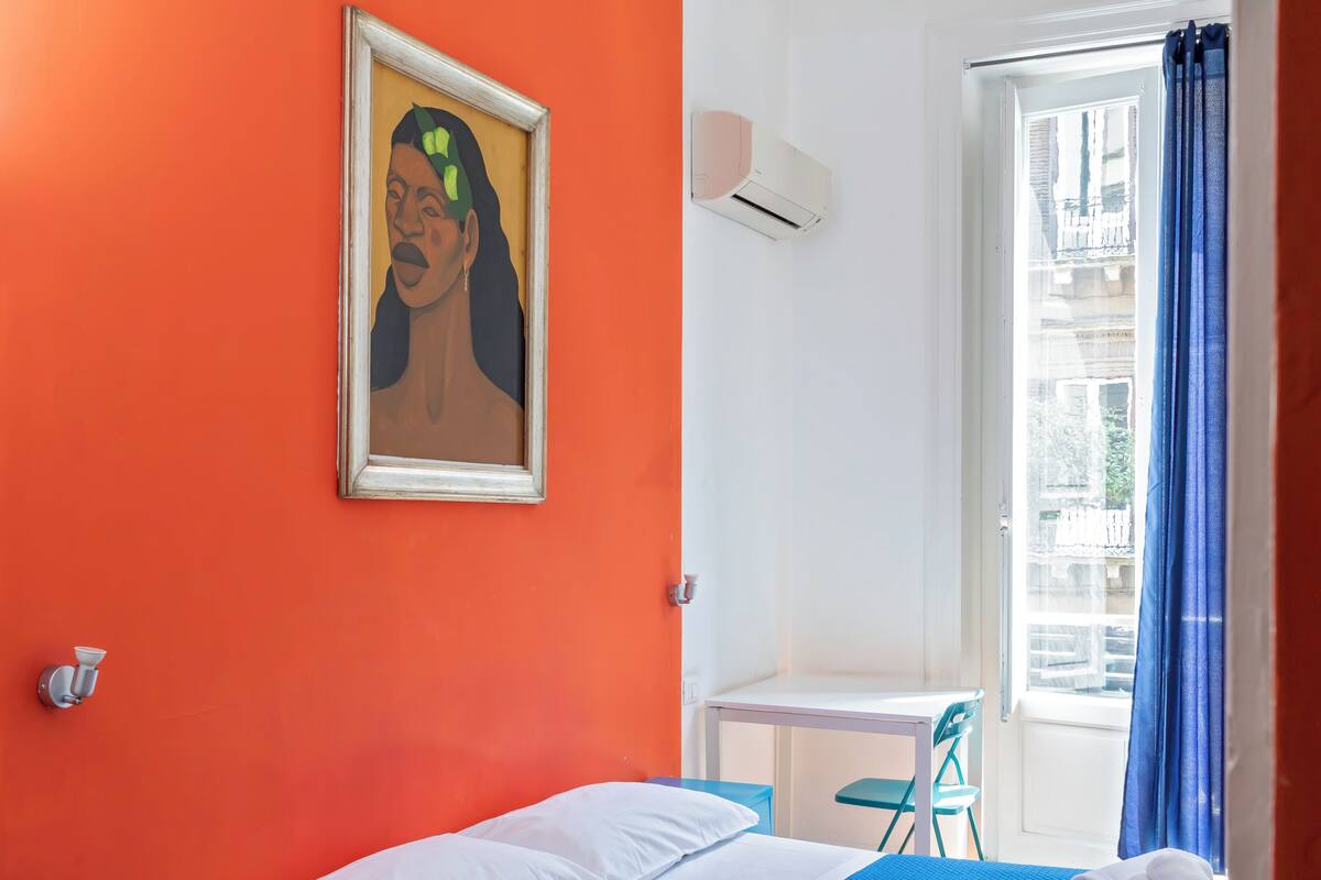 Ein Zimmer mit orangefarbener Wand, einem Bett mit weißer Bettwäsche, einem kleinen Tisch mit Stuhl und einem Fenster mit blauen Vorhängen.