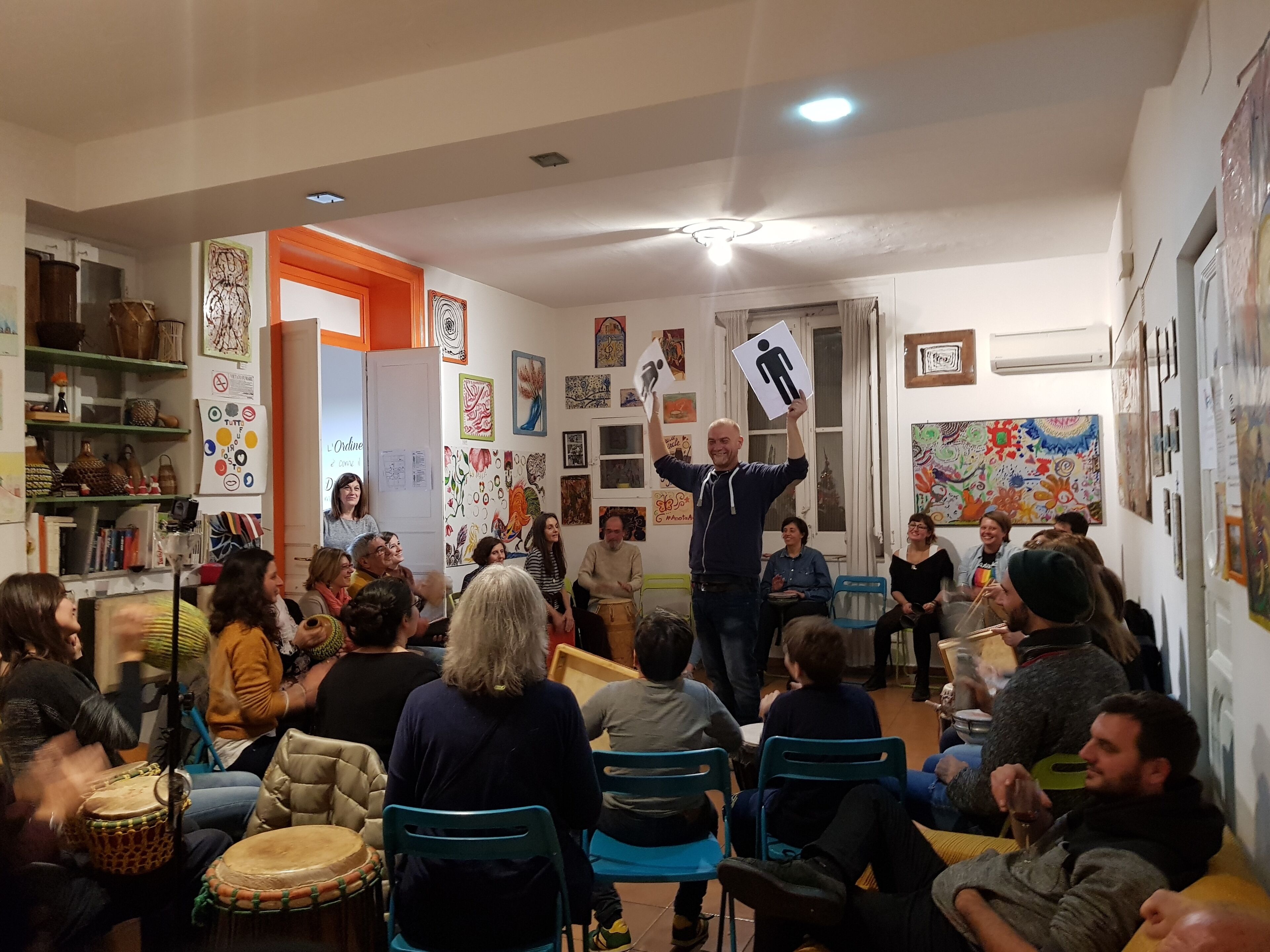 Une pièce où se trouve un groupe de personnes assises et une personne debout, tenant une pancarte. La pièce a un plafond blanc avec un luminaire, une porte avec un cadre orange et des murs décorés de diverses œuvres d’art. Il y a des chaises et une table dans la pièce.