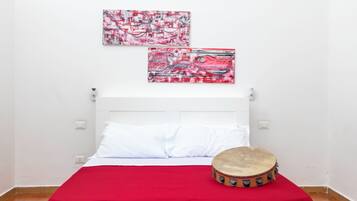 Un lit avec une literie blanche, une couverture rouge et un tambour posé sur le sol.