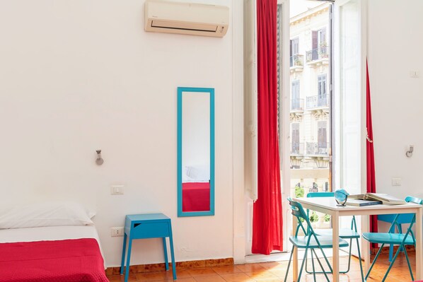A hotel room with a bed, a small table, a chair, a mirror, and a window with red curtains.