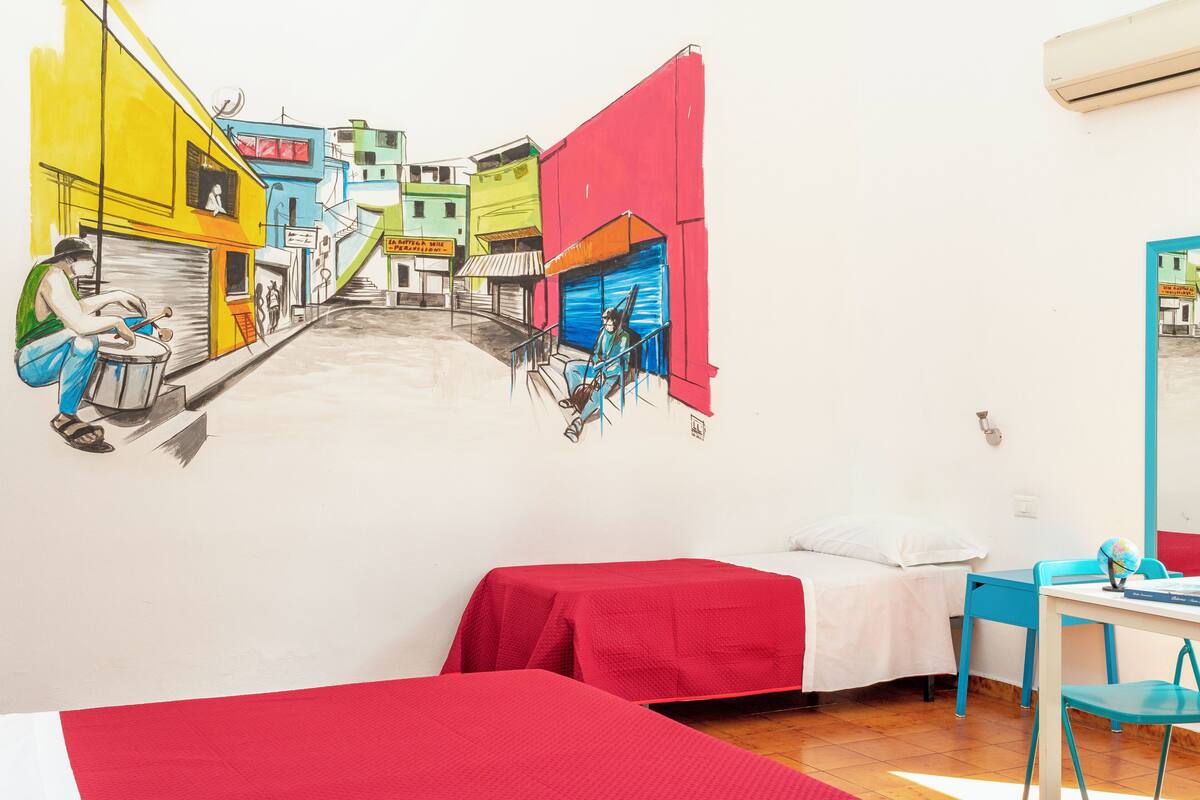 Ein Zimmer mit Wandgemälde einer Straßenszene, einem Bett mit roter Bettdecke, einem blauen Sessel und einem kleinen Tisch mit Globus.