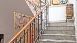 Una escalera de mármol con barandilla de madera y balaustradas de metal, que conduce a una habitación con un mural colorido y un barril rústico.