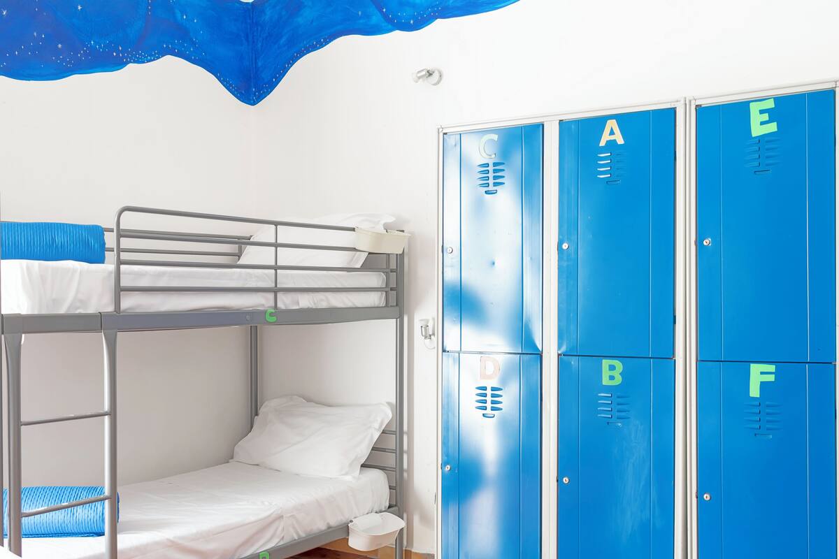 Ein Etagenbett mit weißer Matratze und blauer Decke, ein weißer Eimer und eine Reihe blauer Schließfächer mit Buchstaben.