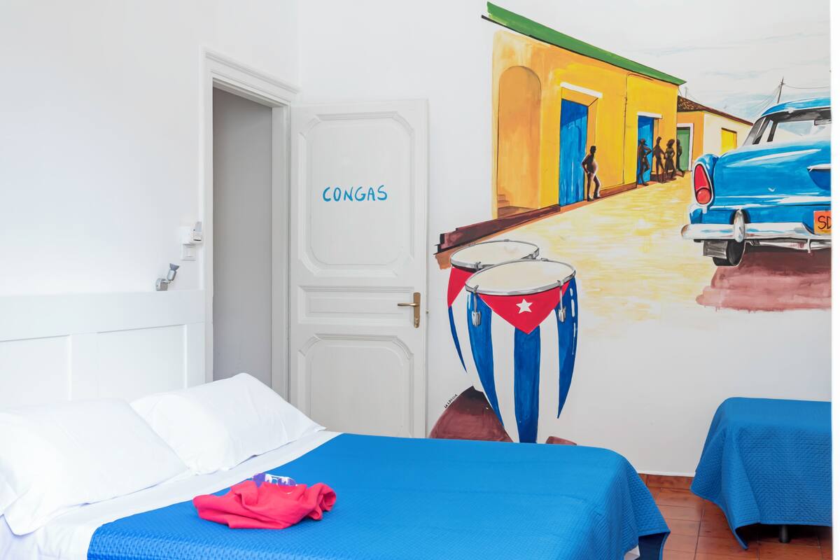 Ein Hotelzimmer mit einem Bett, einer blauen Tagesdecke, einer rot-weißen Trommel und einem Wandgemälde, das eine Straßenszene zeigt.