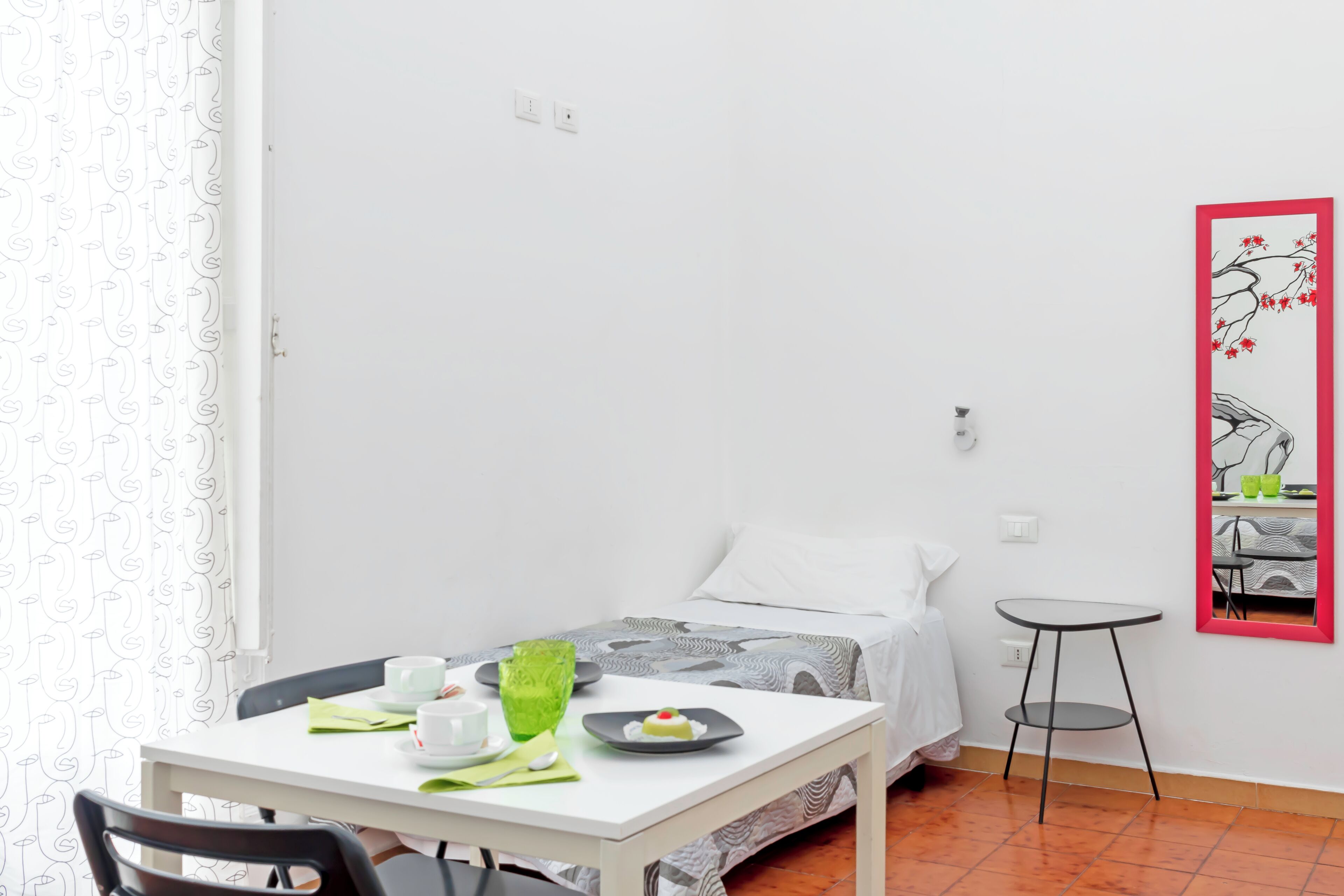 Una habitación pequeña y limpia con una cama, una mesa con un florero verde y un taburete.