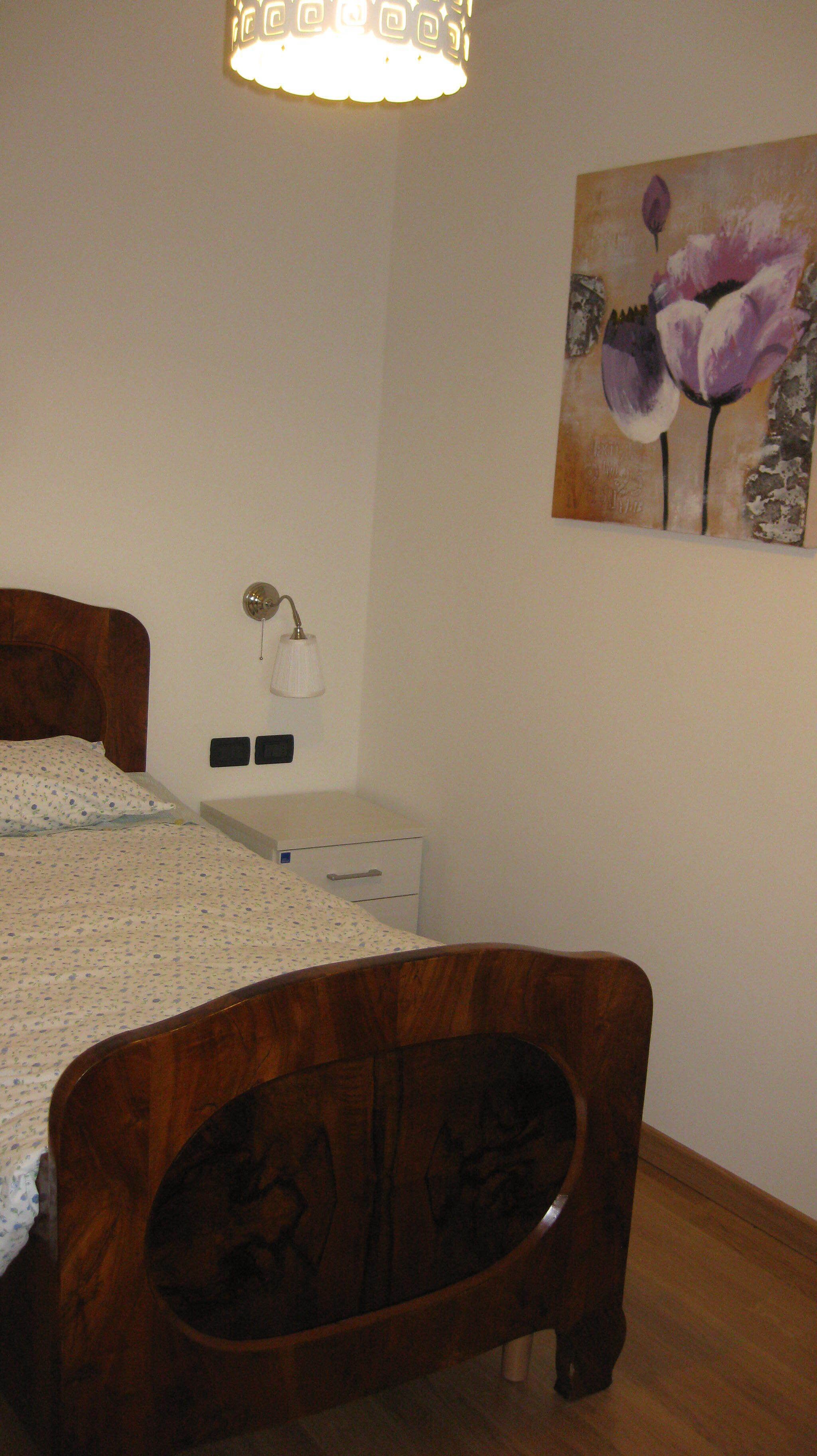 Un dormitorio con cama de madera, una mesita de noche, una lámpara de pared y un cuadro con flores.