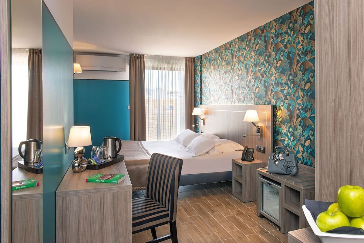 A hotel room with a bed, a desk with a lamp, a chair, a small table with apples, and a decorative wall.