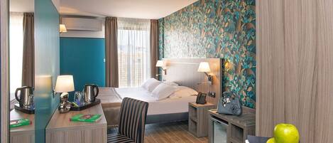 A hotel room with a bed, a desk with a lamp, a chair, a small table with apples, and a decorative wall.