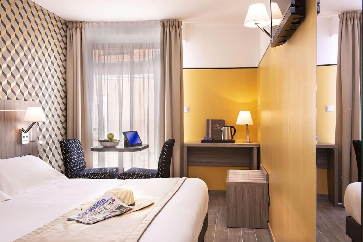 A hotel room with a bed, a desk with a laptop, a chair, a small table with a bowl of fruit, and a lamp.