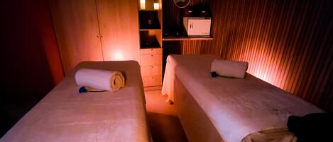 Two single beds with white linens and a small bedside table, a mirror, and a shelf with candles.