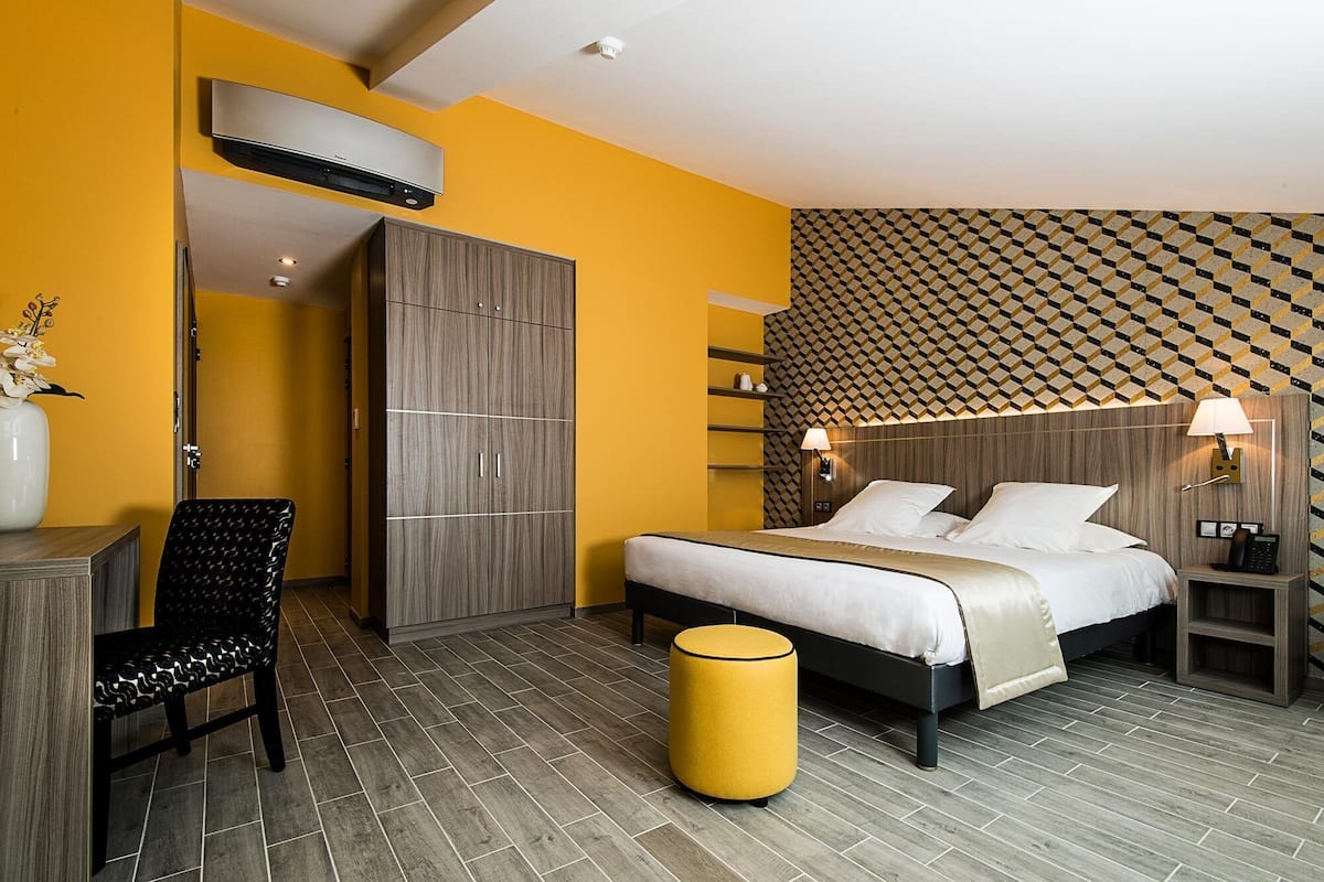 A modern hotel room with a bed, a wardrobe, a chair, a bedside table, a lamp, and a decorative wall pattern.