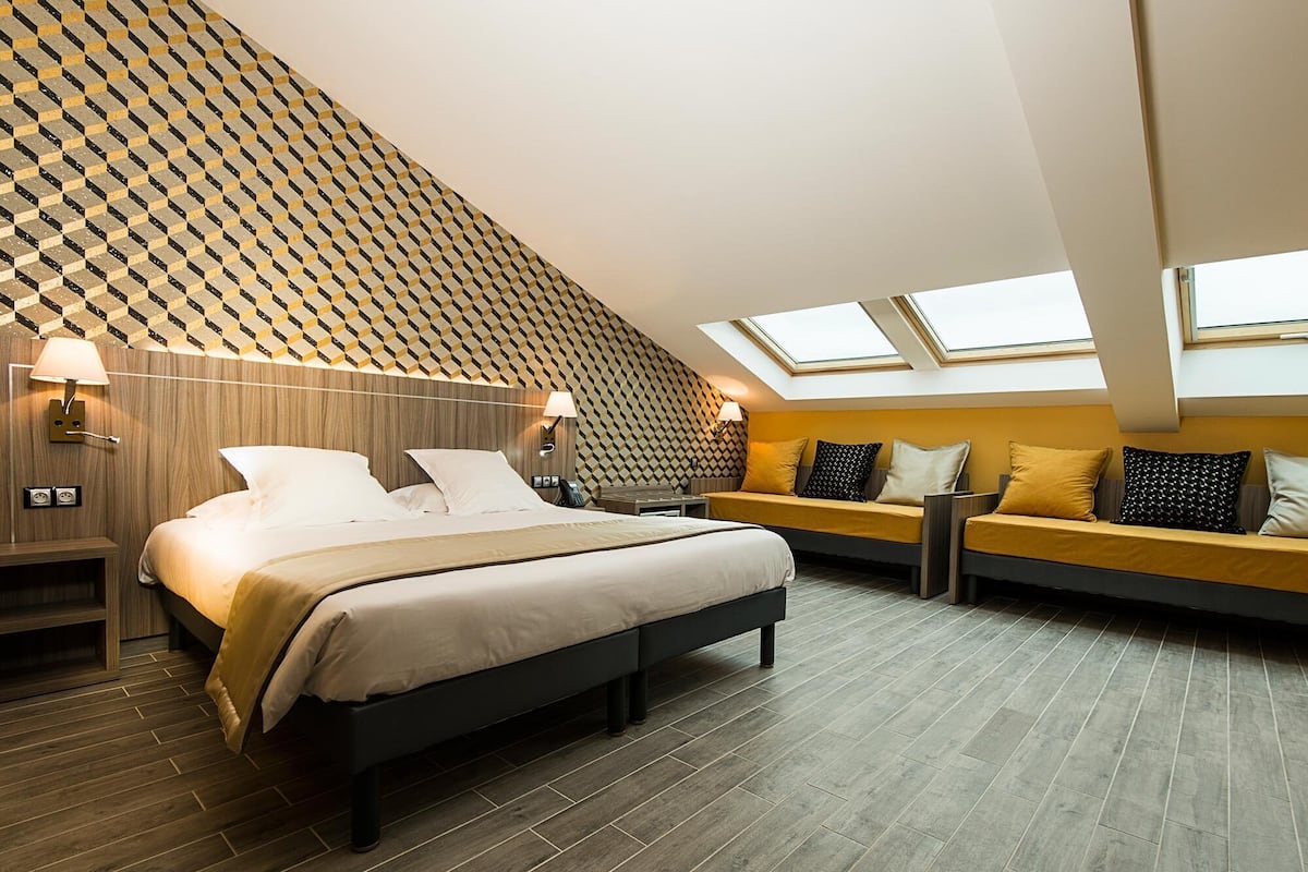 A modern hotel room with a sloped ceiling, a bed, a sofa, and a desk.